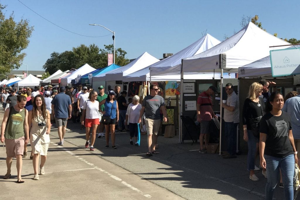 The street is lined with arts & crafts booths. Canopies, mostly white and blue, can be seen stretching off the horizon. A large group of people can be seen going booth to booth.