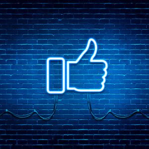 A brightly lit, fluorescent bulb Facebook thumb is lit up blue with light splashing onto the dark brick behind it.