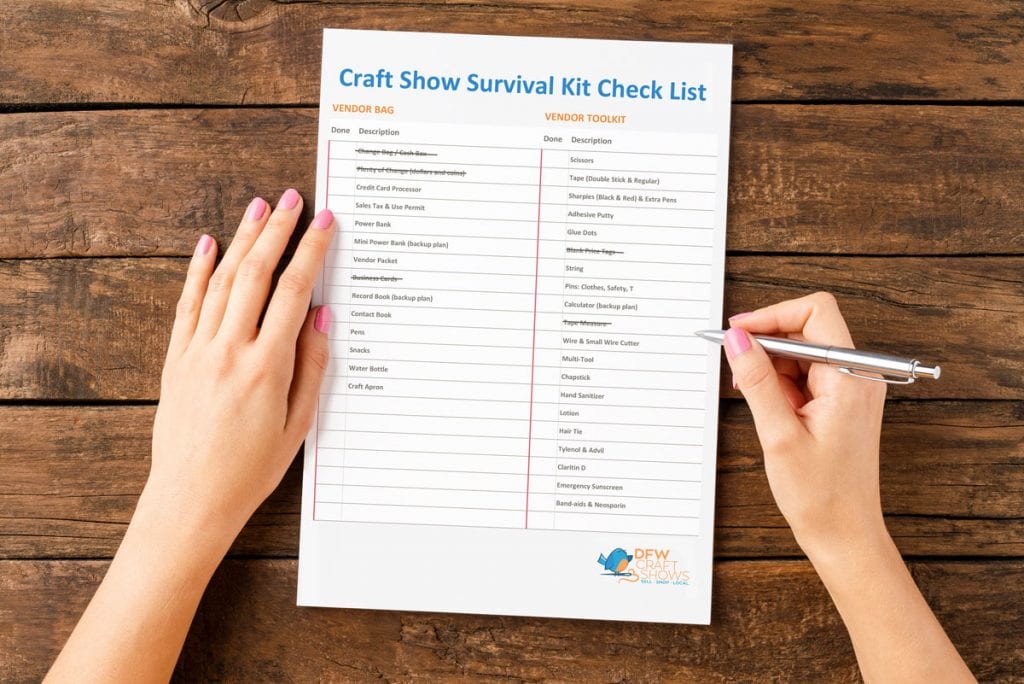 A piece of paper sits on top of a wood table, flanked by two hands with painted nails. One hand holds a pen. The paper reads "Craft Show Survival Kit Checklist