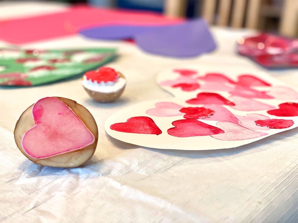 A potato carved into a heart stamp sits on a white table cloth surrounded by painted heart art for valentine's day