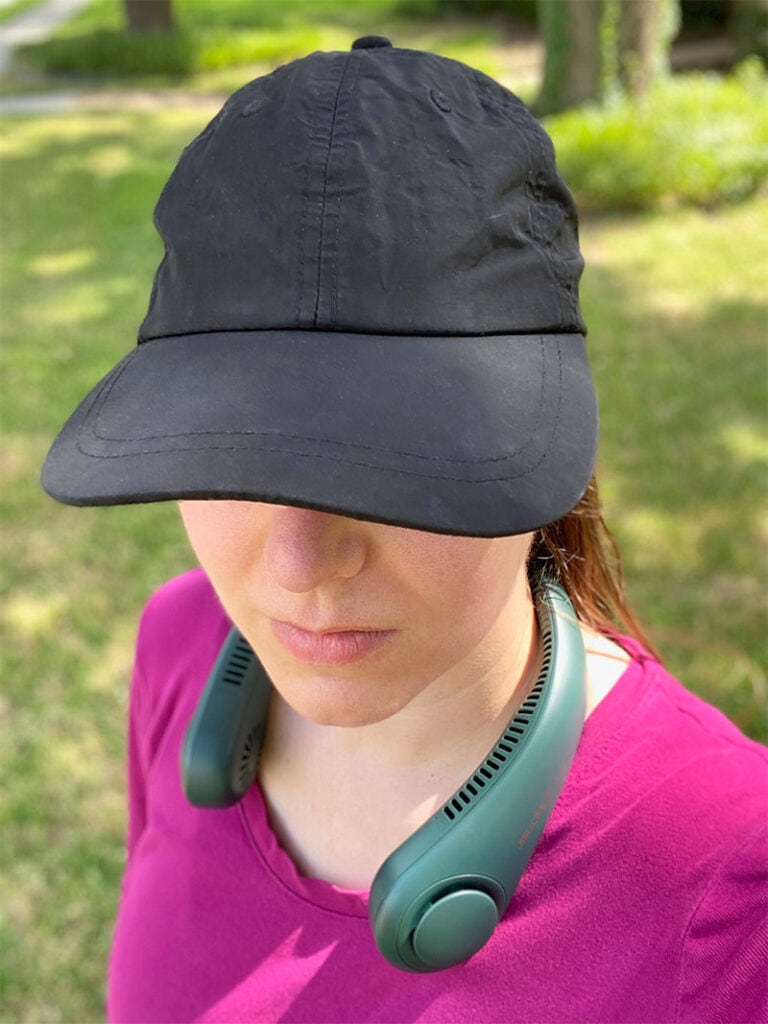 a woman in a purple shirt and black hat wears a personal neck fan outside