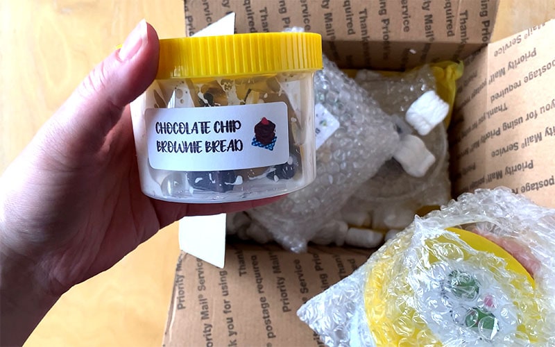 a hand holds a clear slime container with yellow lid above packaging. The label reads "chocolate chip brownie bread"
