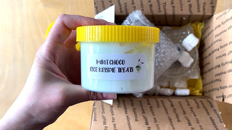 A hand holds a clear slime container with yellow lid that reads "mint choco rice krispie treats"