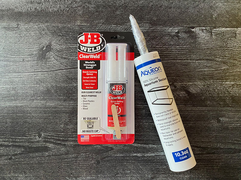 JB Clear Weld Syringe and Aquarium Safe Silicone resting on a dark wood background.
