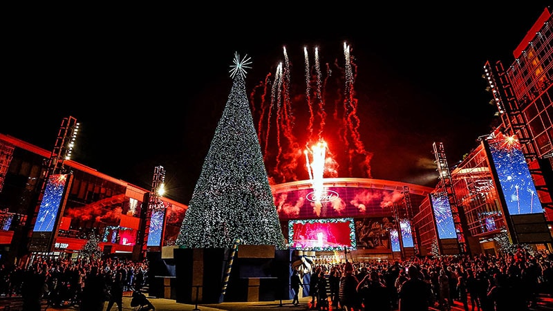 A 67 ft christmas tree is lit with LEDs and stands in the middle of a large plaza with fireworks erupting in the background
