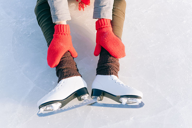 a person sits on the ice with their ice skates on. They have coral color mittens and are wearing a sweater with matching scarf
