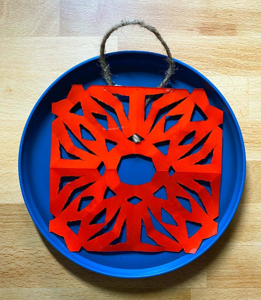 A red paper snowflake sits in a blue plate with twine curled over the top