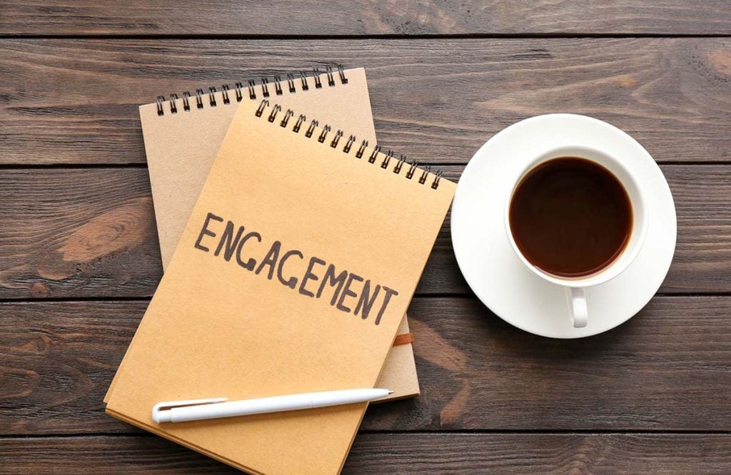 A kraft paper notebook sites on an expresso colored desk next to a cup of coffee, the word "engagement" is written boldly.