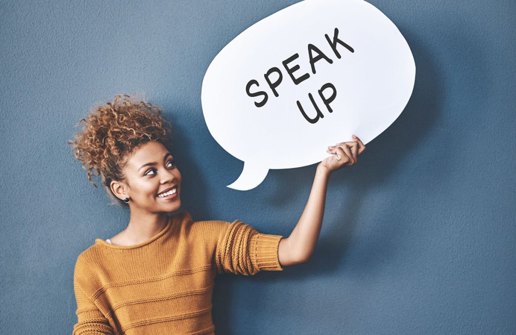 A woman of color holds a large white talk bubble with the words "speak up"