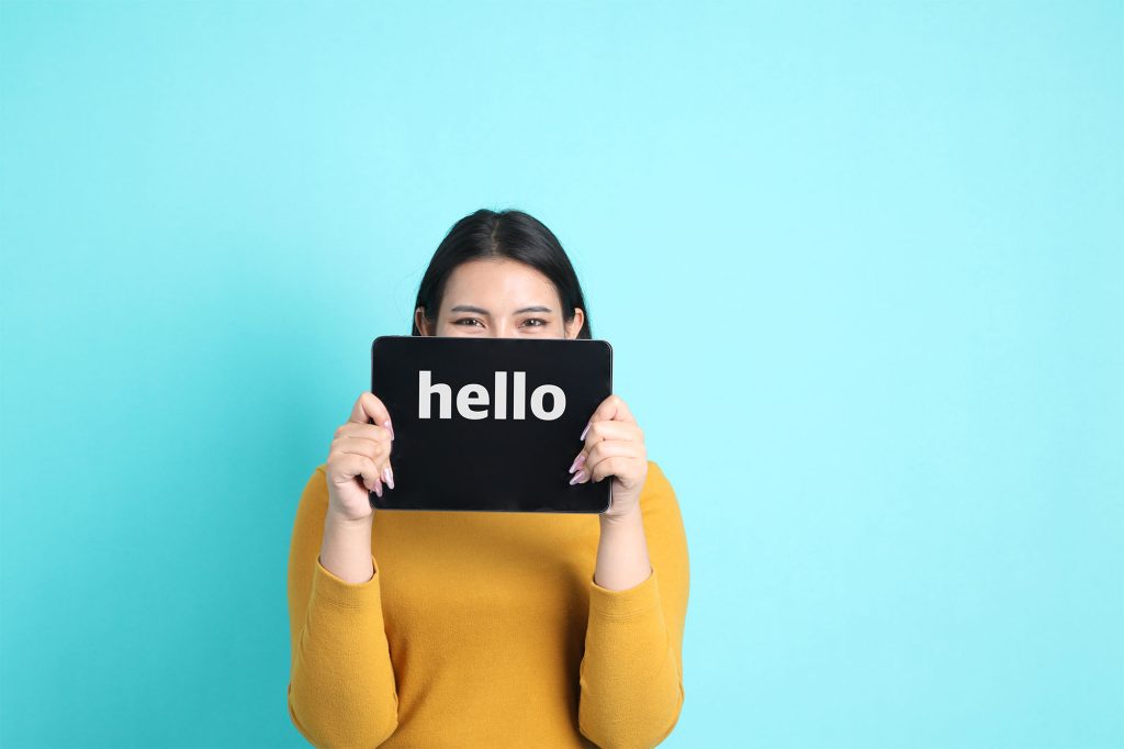 a woman in a yellow shirt stands in front of a blue background. She's partially hiding behind her device, which says hello on the screen.