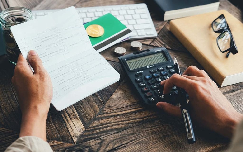 A person sits at a desk with their checkbook and a calculator.  Glasses and a notepad sit nearby.