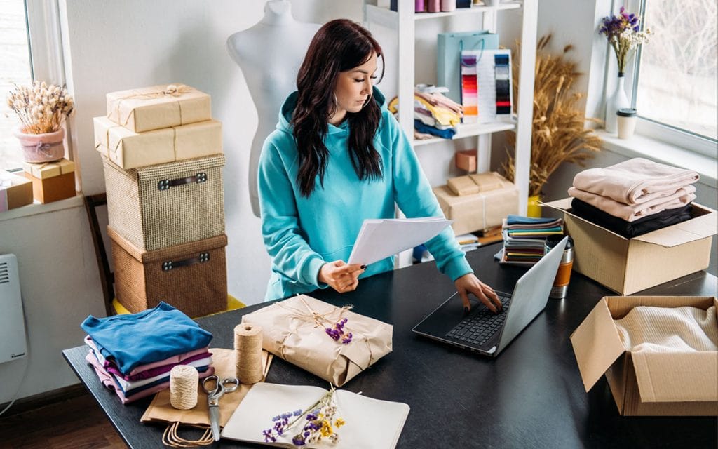 A person in blue stands next to her laptop with several boxes in various states of packaging
