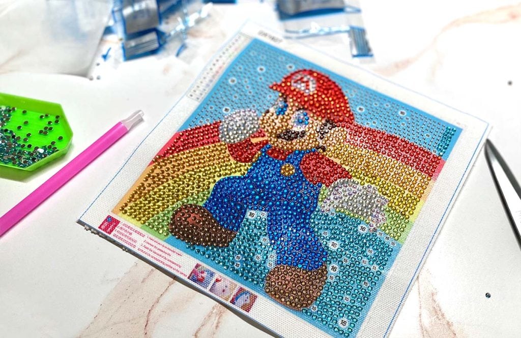 A diamond painting canvas featuring Super Mario with a rainbow background lays on a marble surface with diamond painting tools scattered about