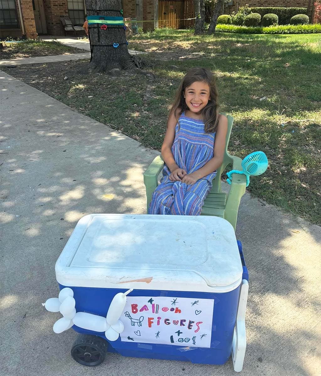 a young girl sits behind an ice chest with a sign that reads "balloon animals $1" with a white balloon animal dog