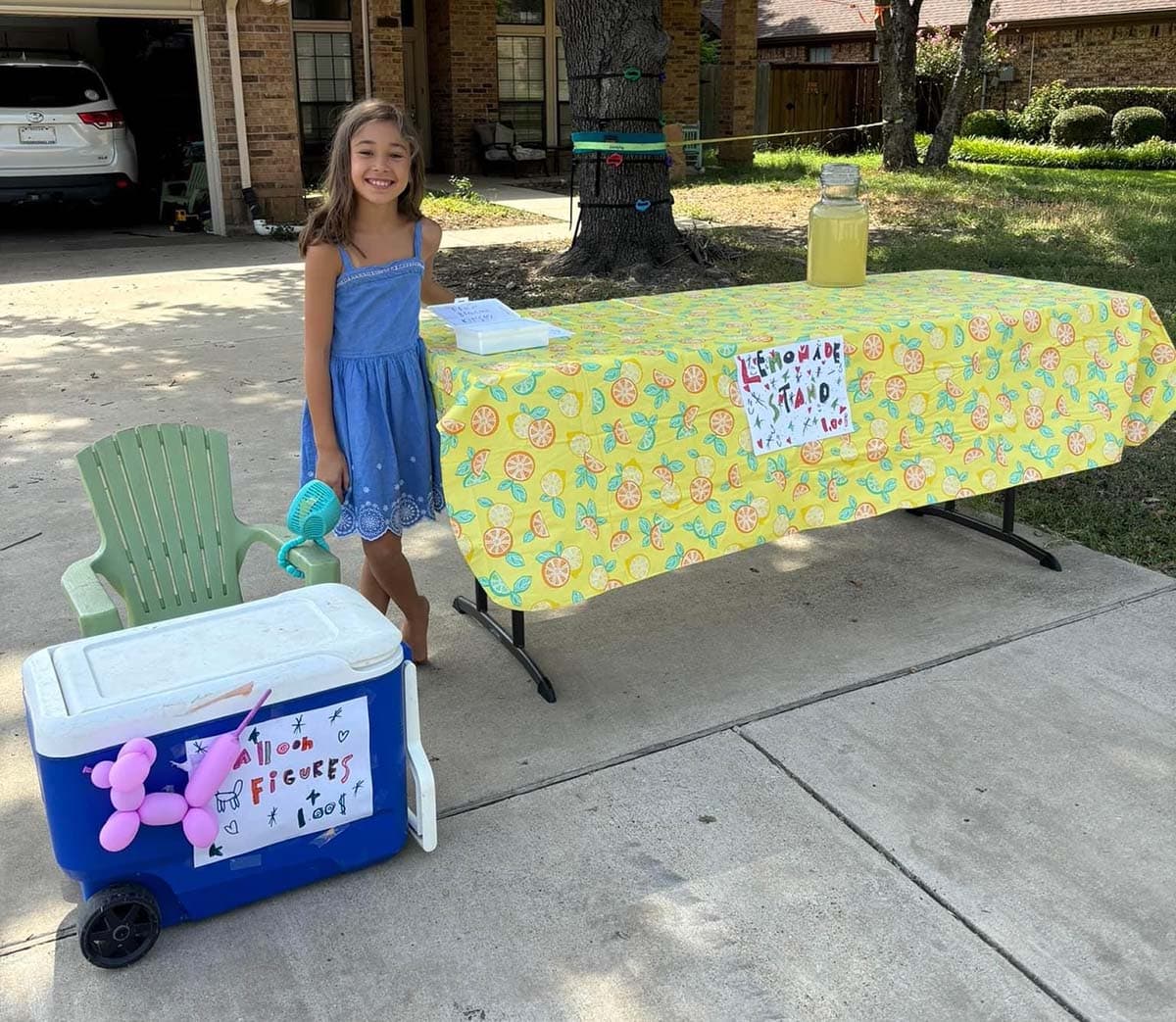 a young girl sits behind an ice chest with a sign that reads "balloon animals $1" with a white balloon animal dog